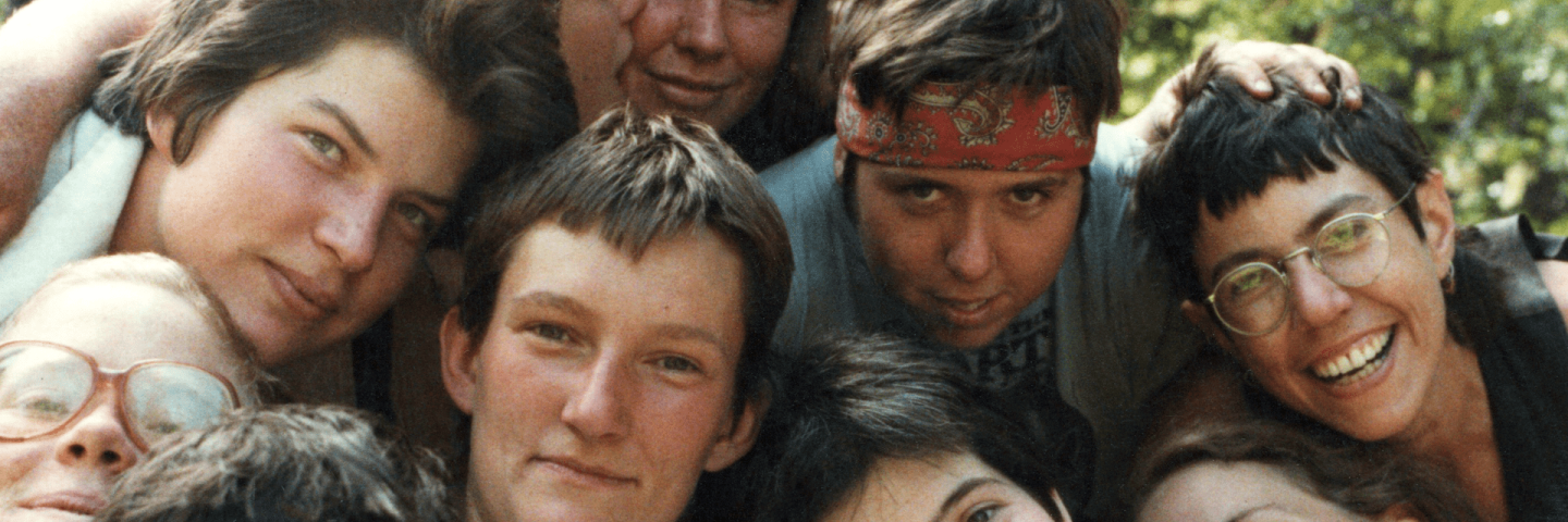 Image of 10 white lesbians in the 1980s clustered very close together, seated, with expressions ranging from joyous laughter to serenity.