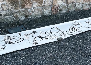 A long scroll of paper on the dark gravel ground. It's held down by normal rocks and is covered in calligraphic swirls of dark ink. 