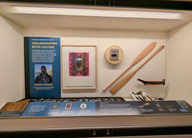 An exhibit case. A panel is on the left with visible text reading "Featured Contemporary Artist: Collaborating with Culture." Inside the case is a framed art piece featuring berries in a mason jar (evoking Andy Warhol's pop art), carved canoe paddles, and a colorful woven blanket. 