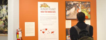 Person stands with back to the camera, looking at a wall with text reading &quot;Hungry Planet: What the World Eats&quot;