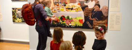 Five small children and an adult holding a baby look at colorful picture of a family gathered around a table.