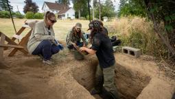 A student stands in a hole and hands something to the students squatting around the whole