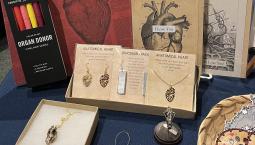 A display of nerdy Valentine's Day related items, including a notebook with an anatomically correct heart on the front, jewelry with skulls and dinosaur tracks, and a candle with a botanical fern print. 