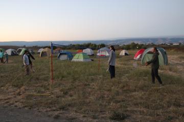 three students play volleyball in front of a backdrop of a prairie with about 10 camping tents visible